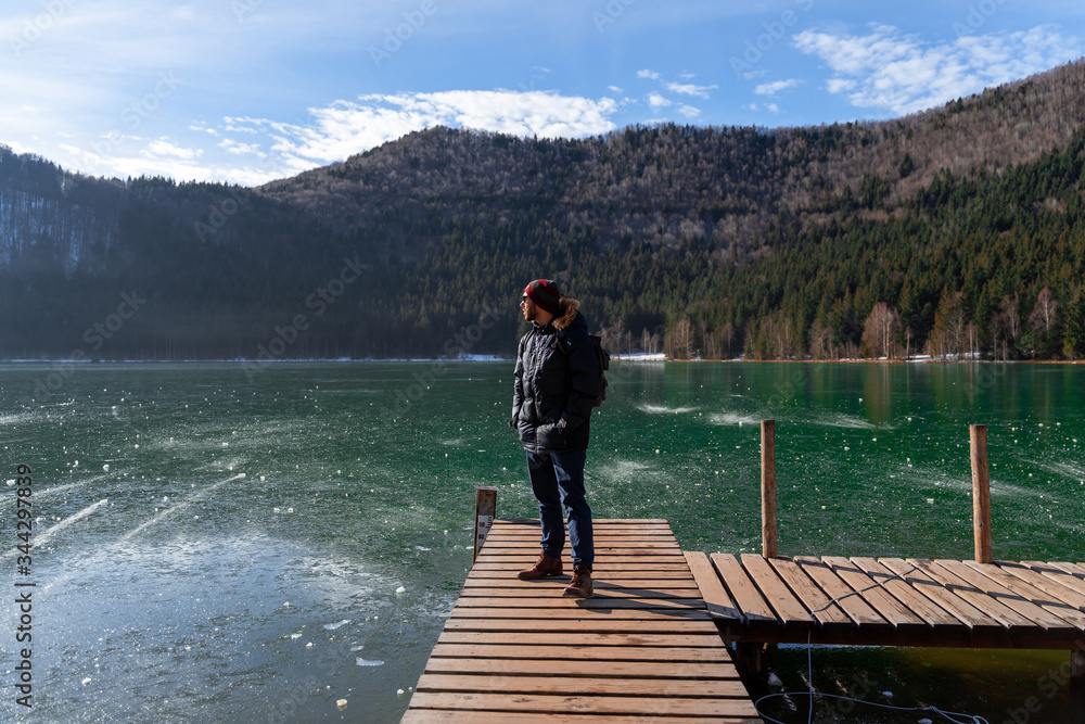 Young male with winter outfit stands looking at frozen lake in Romanian Carpathian Mountains