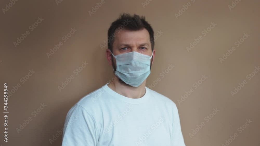 Handsome man in a white T-shirt puts on a medical protective mask and looks at the camera against a beige background. Portrait, the average plan. Natural light, soft focus. Handsome man in a white T-shirt puts on a medical protective mask and looks at the camera against a beige background. Portrait, the average plan. Natural light, soft focus.