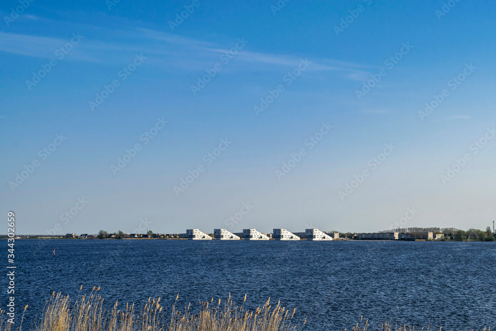 Blue seascape with bright blue water of lake Gooimeer and white modern nautical-style houses on the horizon. Interesting attractions for domestic vacation tourism in Netherlands, Dutch town Huizen. 