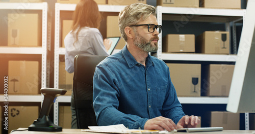 Canvas Print Caucasian gray-haired man in glasses working at computer screen in postal delivery store and typing on keyboard