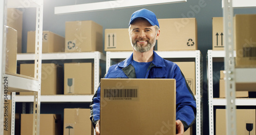 Portrait of happy delivery man in uniform and cap smiling cheerfully to camera with carton box. © VAKSMANV