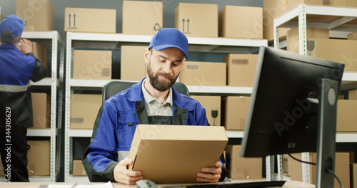 Portrait of delivery man in uniform working at computer in post office store with parcels. Postman scanning bar code on carton box with scanner and registering it on computer.