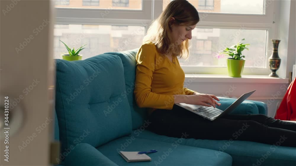 Нoung woman in yellow clothes sits on large blue sofa and types text on laptop keyboard. Girl with curly hair in spacious room with large windows uses a laptop. Remote work during quarantine. Нoung woman in yellow clothes sits on large blue sofa and types text on laptop keyboard. Girl with curly hair in spacious room with large windows uses a laptop. Remote work during quarantine.