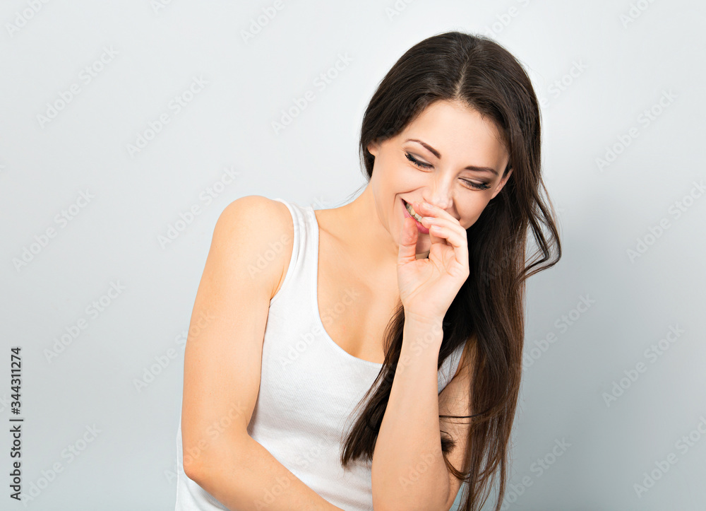 Beautiful sporty chuckling woman covering the hands the face in casual white t-shirt and long hair on blue background. Closeup Beautiful sporty chuckling woman covering the hands the face in casual white t-shirt and long hair on blue background. Closeup