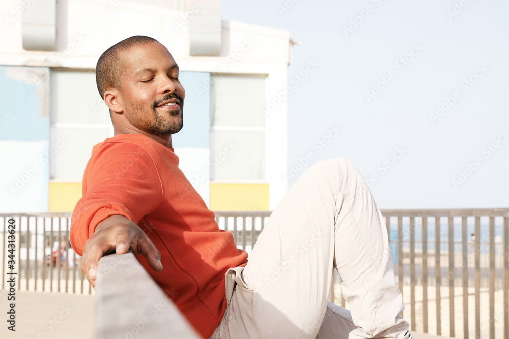 Happy African American man sitting on bench in park wearing casual clothing. Horizontal.
