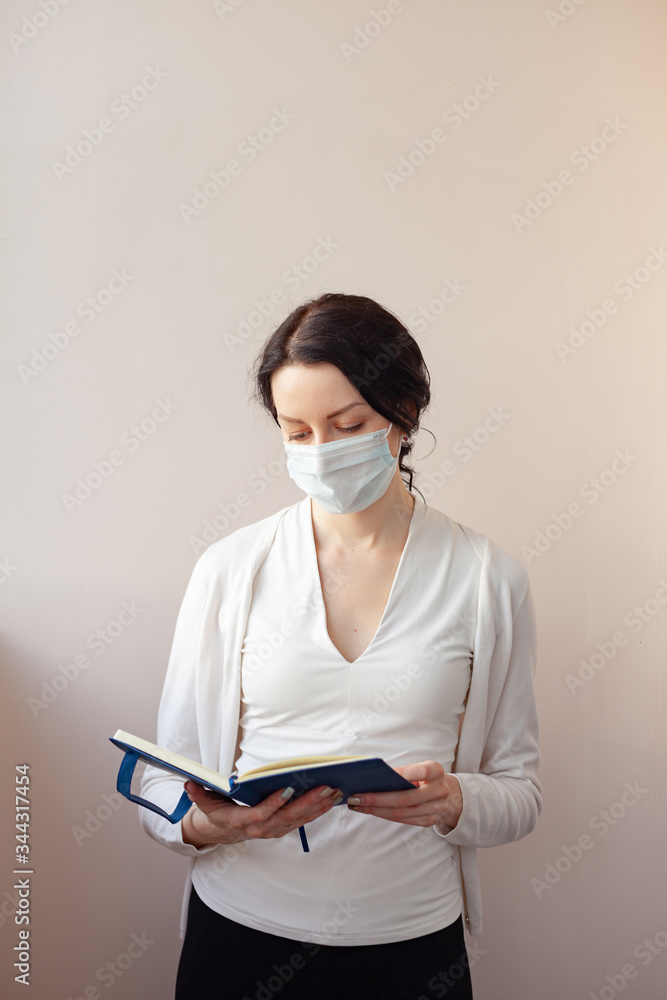A woman in a protective mask stands on a light background in a white jacket and holds a diary in her hands. A medical professional who takes notes