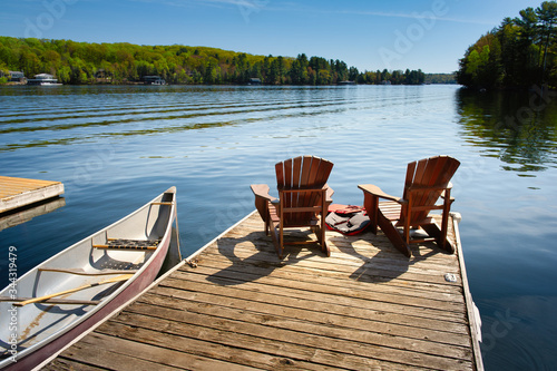 Fotografia Two Adirondack chairs on a wooden dock facing the blue water of a lake in Muskoka, Ontario Canada