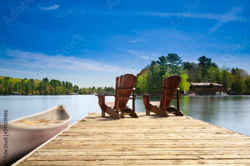 Two Adirondack chairs sit on a wooden dock facing the blue waters of a calm lake. A canoe is tied to the dock. In the background there's a brown cottage nestled between trees. 