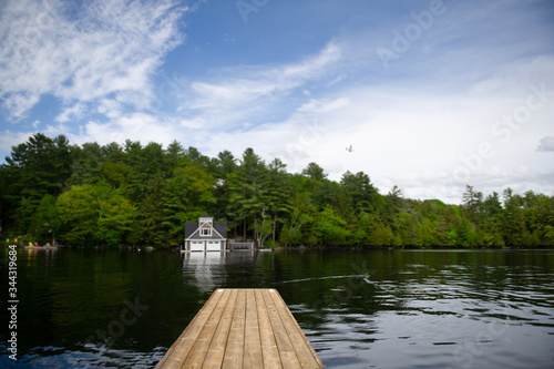Fototapeta Naklejka Na Ścianę i Meble -  Wooden dock on a calm lake in Ontario, Canada. A Boat house, nestled between green trees, is visible across the water. A plane is crossing the blue sky.