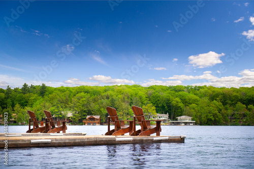 Wallpaper Mural Muskoka chairs on a wooden dock overlooking a calm lake. Cottages nestled between green trees are visible across the blue water. Torontodigital.ca