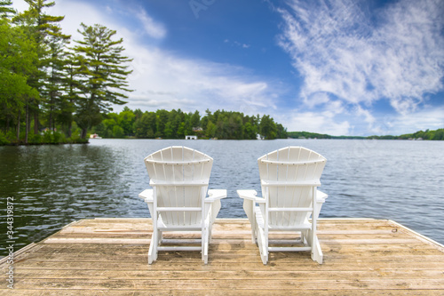 Two white Muskoka chairs on a dock overlooking a lake in Ontario Canada. Across the calm water there's a white cottage nestled between green trees.