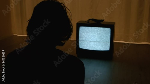 Teenager with disheveled haircut is staring into broken TV sitting on the floor in a dark room, lit by the flickering screen. The handheld camera from behind teenager shoulder.