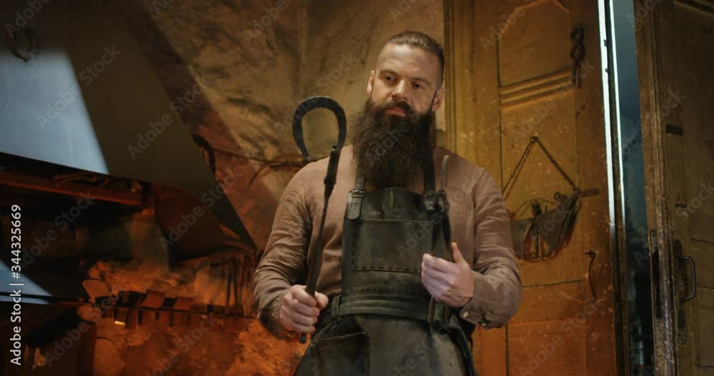 Portrait shot of Caucasian brutal male smith with long beard posing to camera with smile and demonstrating handmade metal horseshoe in smithy. Handsome man smiling to camera with steel items in forge. Portrait shot of Caucasian brutal male smith with long beard posing to camera with smile and demonstrating handmade metal horseshoe in smithy. Handsome man smiling to camera with steel items in forge.