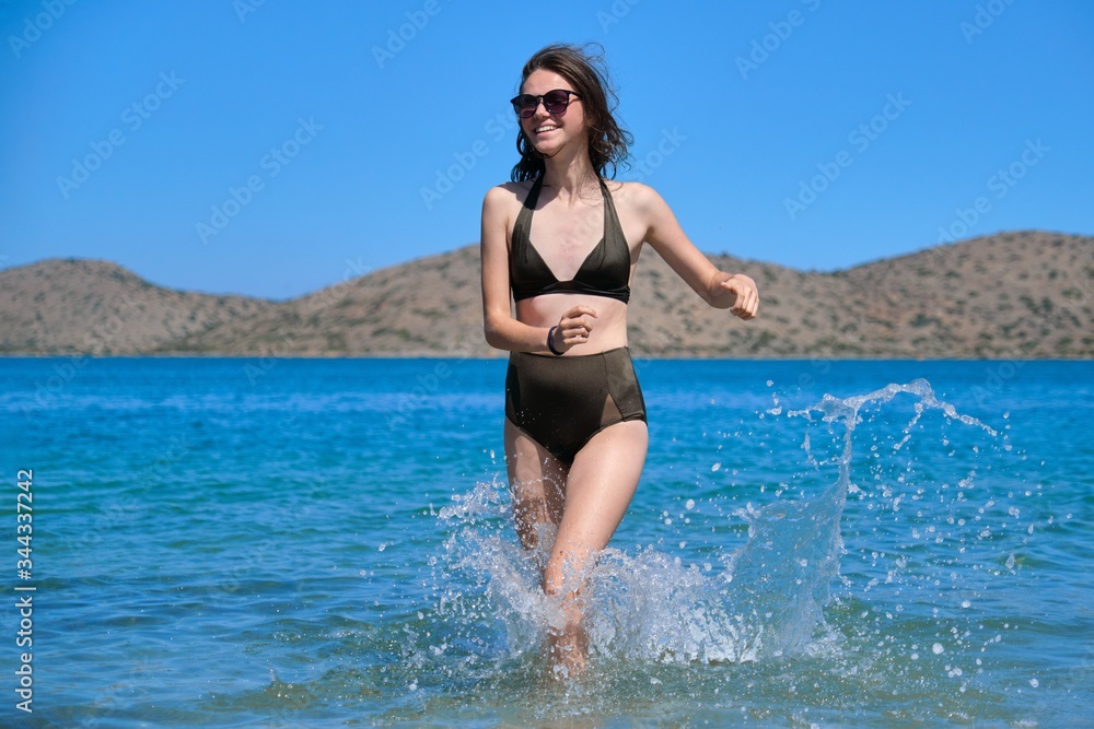 Beautiful laughing teenager girl in sunglasses swimsuit bikini running on sea water Beautiful laughing teenager girl in sunglasses swimsuit bikini running on sea water