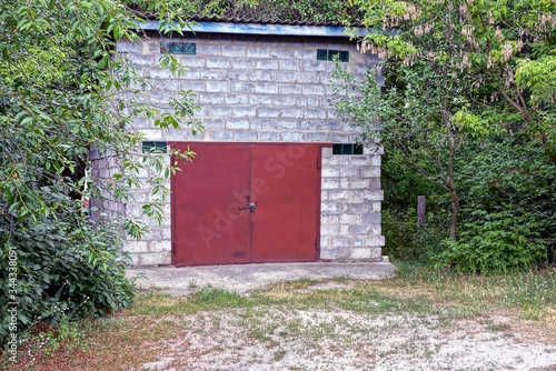 one white cinnamon garage with closed red iron gates among greenery and trees outside