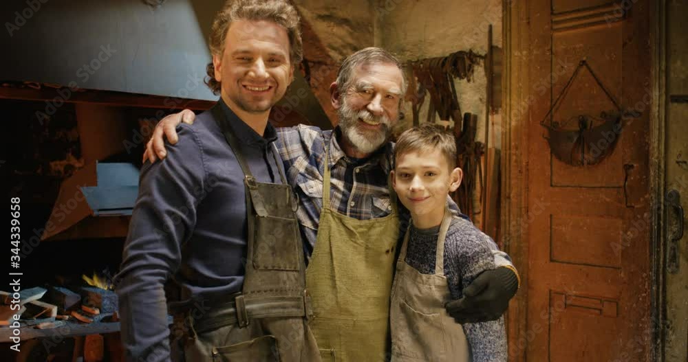 Portrait shot of happy joyful Caucasian old man with son and grandson hugging and smiling to camera in smithy worksop. Male family of forge craftsmen. Three generations of smiths. Portrait shot of happy joyful Caucasian old man with son and grandson hugging and smiling to camera in smithy worksop. Male family of forge craftsmen. Three generations of smiths.