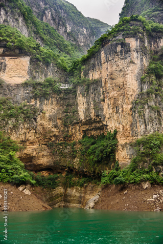 Wuchan  China - May 7  2010  Dragon Gate Gorge on Daning River. Portrait of tall brown and black cliffs descending into emerald green water. Green foliage on top and bottom. Silver sky.