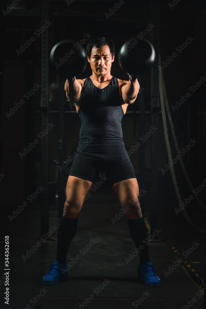 asian athletic strong man having workout and bodybuilding with kettlebell weightlifting backsquat style in gym and fitness center in dark tone
