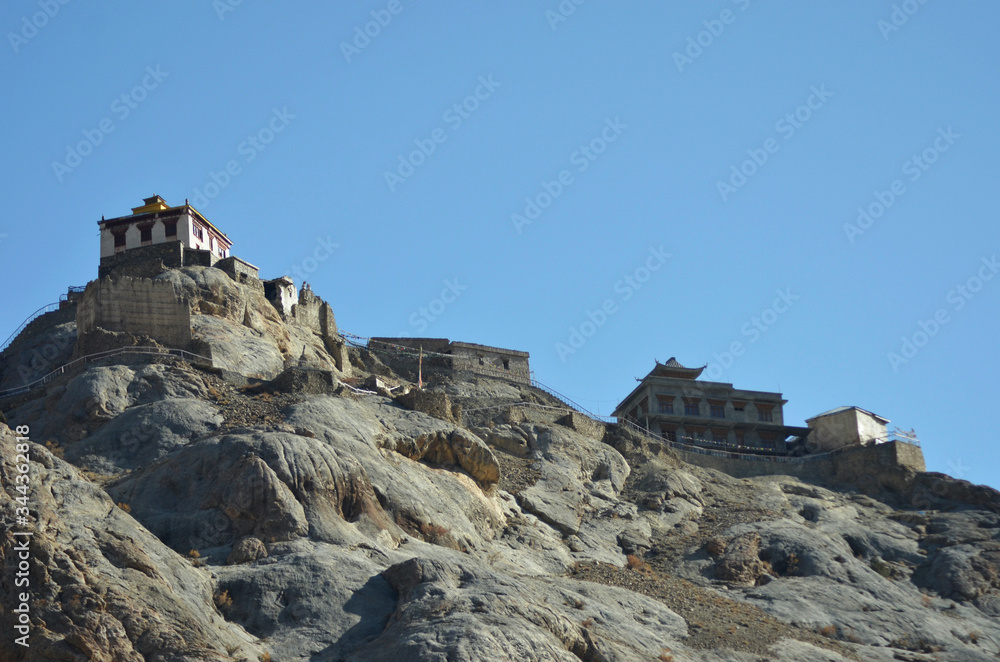 White monastery buildings are seen against a clear blue sky in Ladakh, India. Prayer flags are hung along a path on top of the rocky hill leading to the Buddhist monastery.