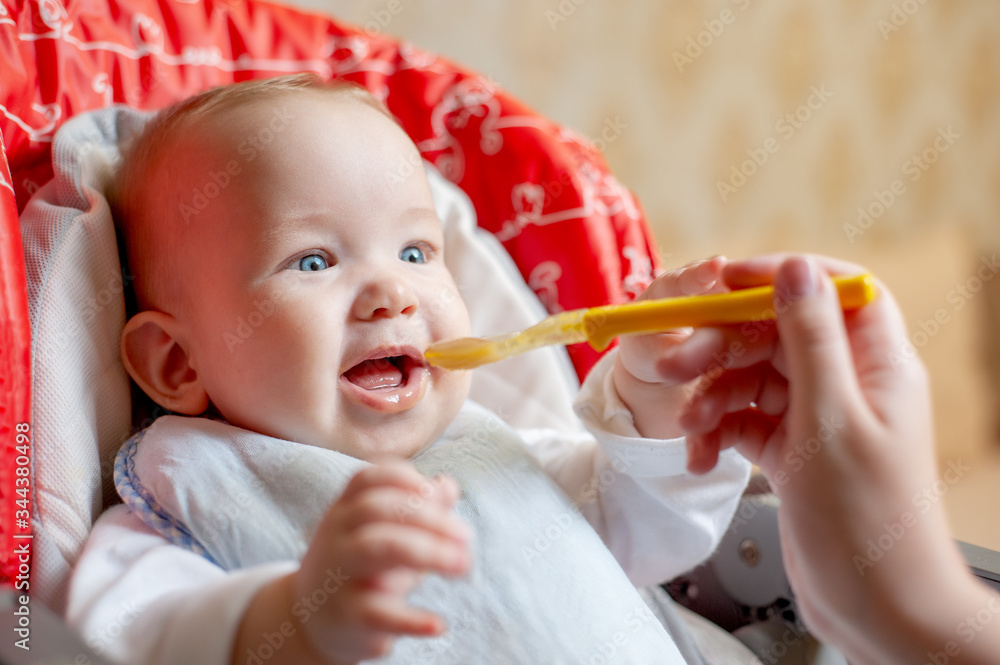Mother feeding her happy baby son with spoon. Mother giving healthy food to her adorable child at home. Parent feeding baby boy in high chair. Close-up Mother feeding her happy baby son with spoon. Mother giving healthy food to her adorable child at home. Parent feeding baby boy in high chair. Close-up