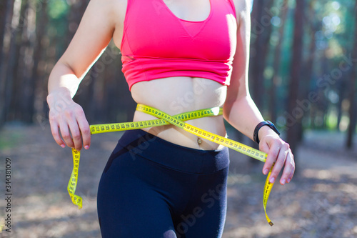 Photo of a fit and healthy young lady measuring her waist with a tape measure controlling her weight loss while training in the forest
