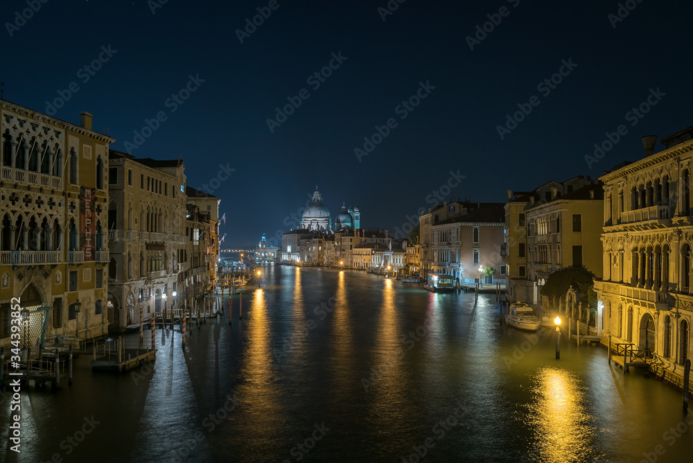 A scenic view of a beautiful Venetian canal at night with colourful architecture, a bridgeand reflections of boats running along the water in the town of Venice, Italy