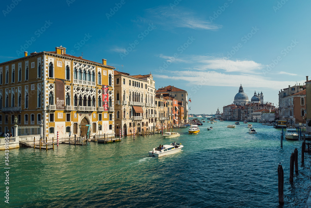 A scenic view looking down one of the main Venetian canals on a warm sunny day. This photo shows colourful houses and boats cruising along the beautiful blue water in the town of Venice, Italy