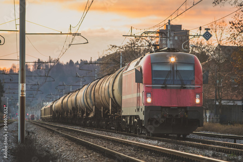 European style freight train with a modern locomotive on the front, carrying liquids and other goods on a straight track in an urban setting during evening hours.