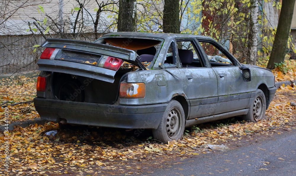 Old broken abandoned dark gray car on yellow leaves in the yard of an apartment building