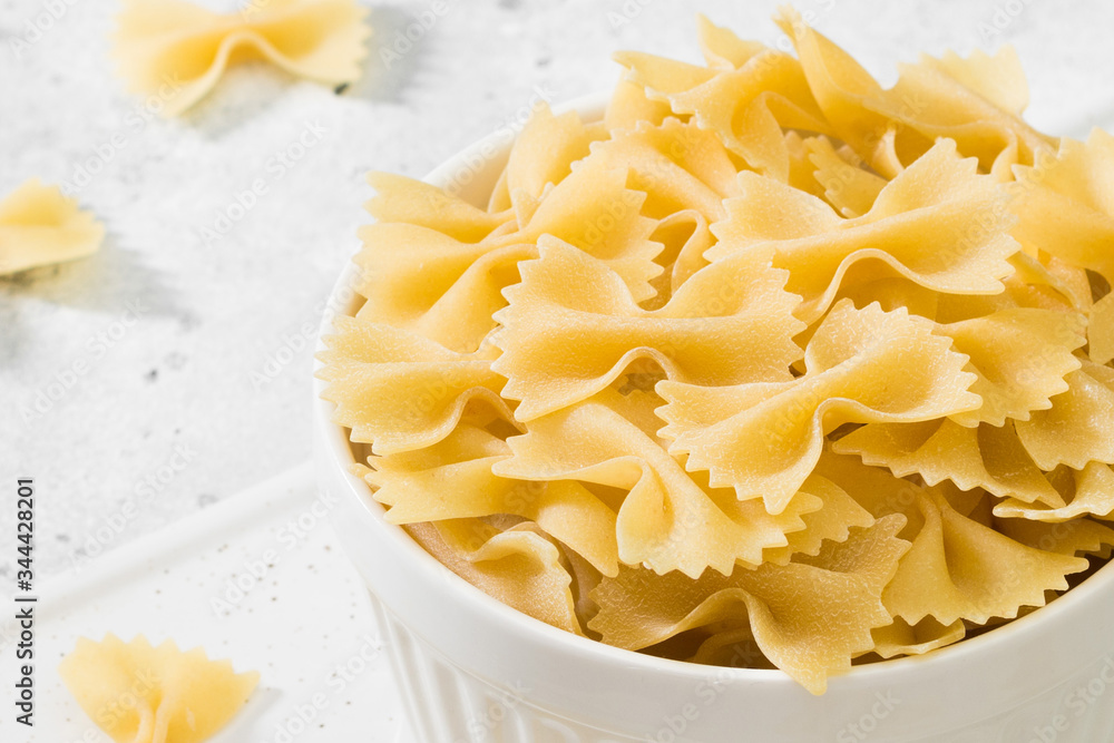 Raw Farfalle pasta in a white bowl on a light gray table. Farfalle macaroni close up with space for text Raw Farfalle pasta in a white bowl on a light gray table. Farfalle macaroni close up with space for text