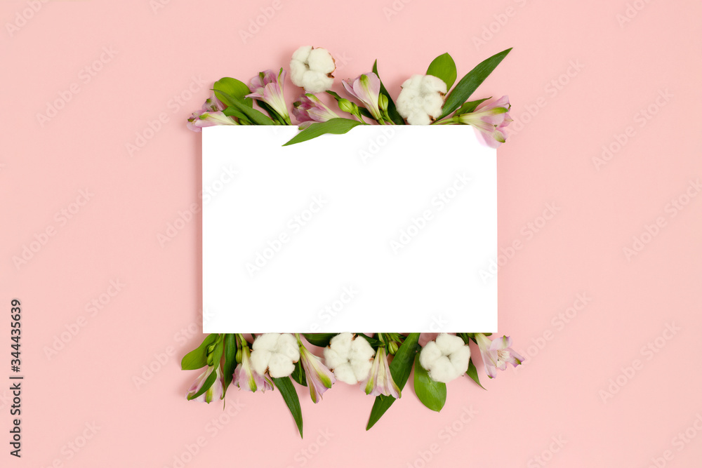 Paper card mockup with frame made of green leaves, cotton, alstroemeria flowers on a pink pastel background. Holiday floral concept.
