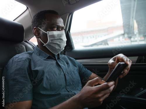 Side view of one black businessman wear mask traveling in the car using mobile phone looking at phone in hand during pandemic of coronavirus Covid-19