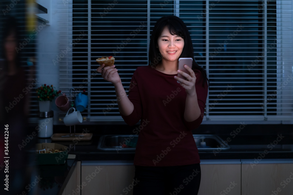 Asian woman virtual happy hour meeting and eating delivery pizza from the box online with friend or taking photo using mobile phone camera in kitchen at night during time of home isolation..
