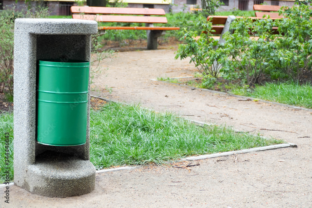a green metal urn in a concrete structure for garbage in a Park among a wooden brown bench and greenery a green metal urn in a concrete structure for garbage in a Park among a wooden brown bench and greenery