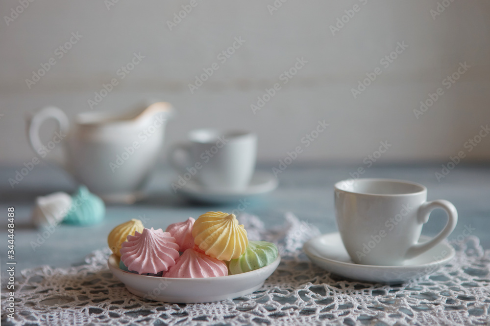 Multi-colored meringues in pastel shades with cups and a jug on a light textured wooden background. The concept of Breakfast with tea or coffee, home comfort and tenderness. Close-up, selective focus Multi-colored meringues in pastel shades with cups and a jug on a light textured wooden background. The concept of Breakfast with tea or coffee, home comfort and tenderness. Close-up, selective focus