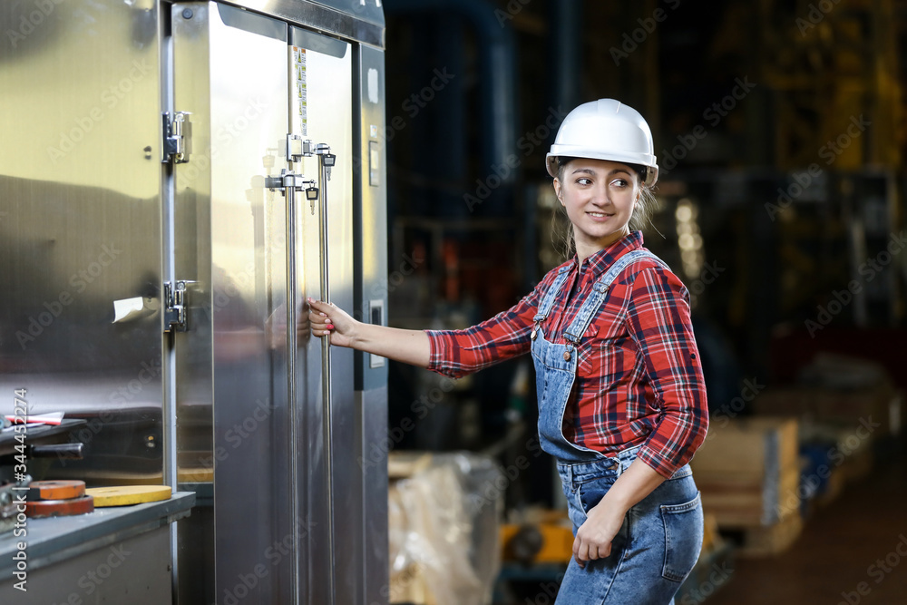 Young girl in a work dress and white hard hat in a factory. Woman in a work uniform. Working process.
