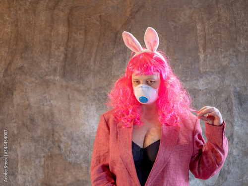 Attractive girl in pink wig and medical mask in plaid shirt, pink jacket and carnival ears of hare. Beautiful middle aged woman with cancer without hair