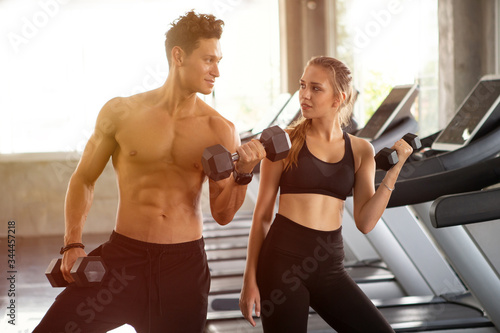 Fototapeta Naklejka Na Ścianę i Meble -  young fitness couple lifting dumbbells together at  gym in morning light .  bodybuilder man and woman showing muscle . two people exercise or workout in sport club. training healthy lifestyle .