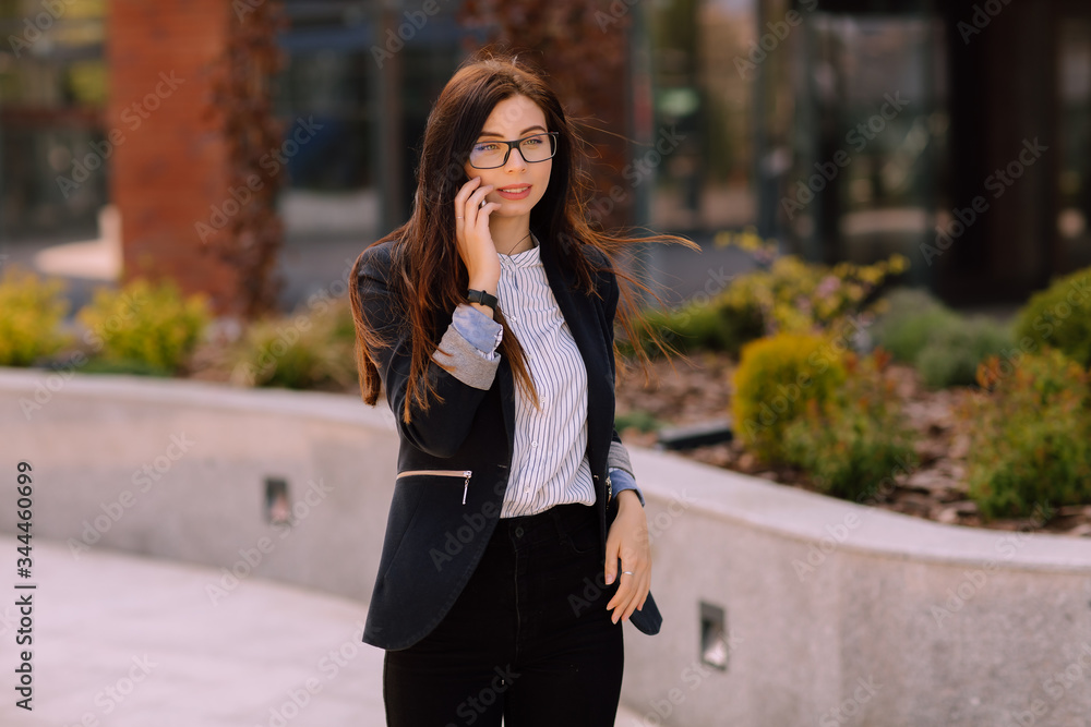 Young successful business woman with long brunette hair in glasses and suit talking on a mobile phone near office building. 