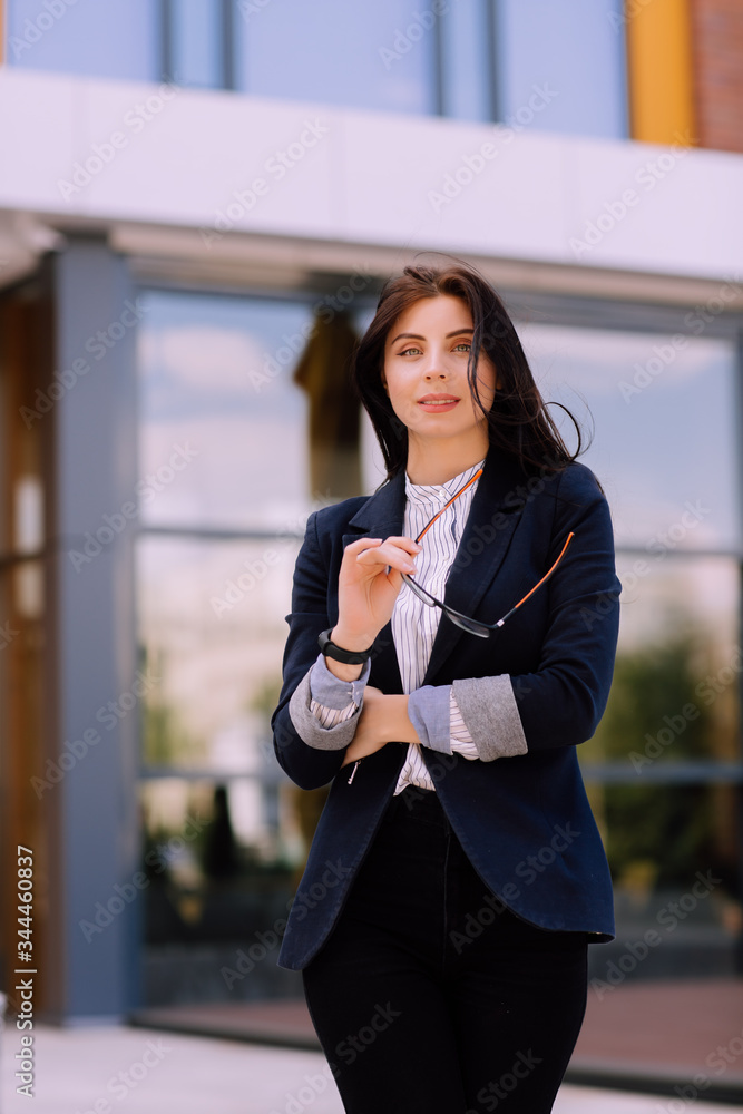 Young successful business woman with long brunette hair in suit stay with arm crossed and hold her glasses near office building. Young successful business woman with long brunette hair in suit stay with arm crossed and hold her glasses near office building.
