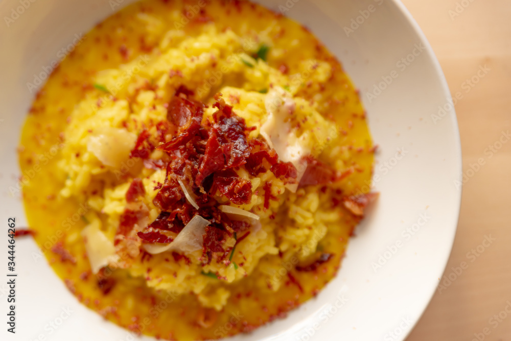 saffron risotto close up, classic yellow colorful italian food staple dish on white plate on wooden table with speck, a gourmet sophisticated presentation