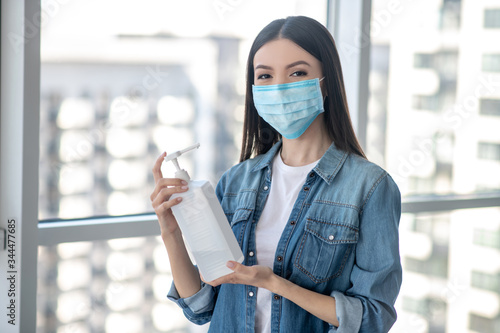 Dark-haired woman in a facial mask holding a bottle with sanitizer for home