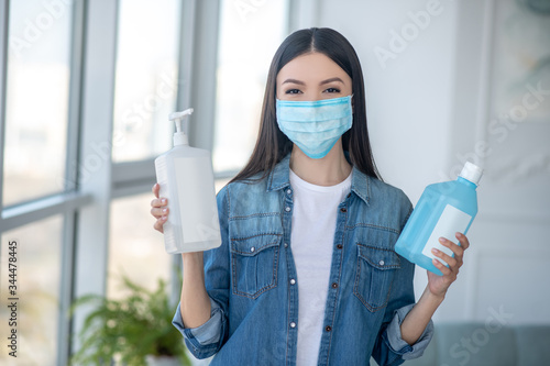 Dark-haired woman in a facial mask holding bottles with disinfecting liquid photo