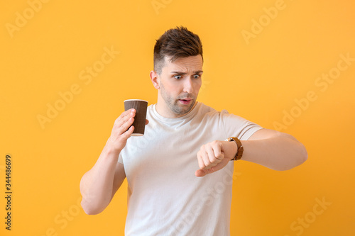 Worried young man with coffee looking at watch against color background