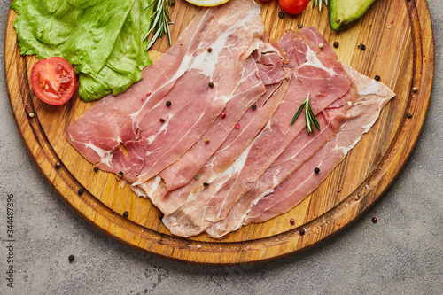 Italian prosciutto crudo or spanish jamon. Raw ham on stone cutting board decoreted with spices, lettuce, lemon, tomatoes and avocado