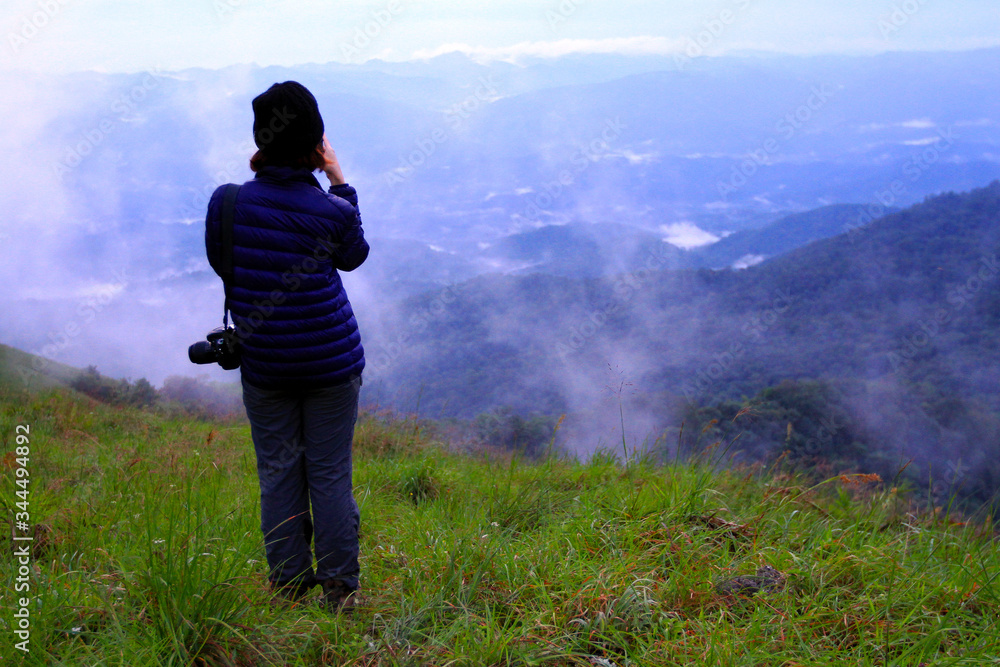 Asian hiking or tourist woman in dark blue sweater with camera standing on green grass field with mountain view and mist background and copy space. Beautiful in nature and Landscape