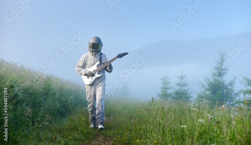 Cosmonaut wearing white space suit and helmet holding white guitar is walking along green mountain meadow in the foggy morning, concept of discovering Earth, music, astronautics and nature.