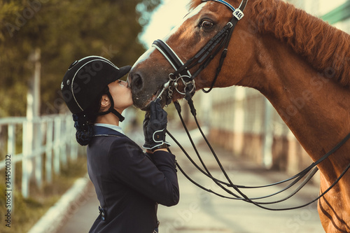 Young teenage girl equestrian kissing her favorite red horse. Multicolored outdoors horizontal image. Dressage outfit 