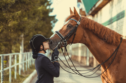 Young teenage girl equestrian kissing her favorite red horse. Multicolored outdoors horizontal image. Dressage outfit 