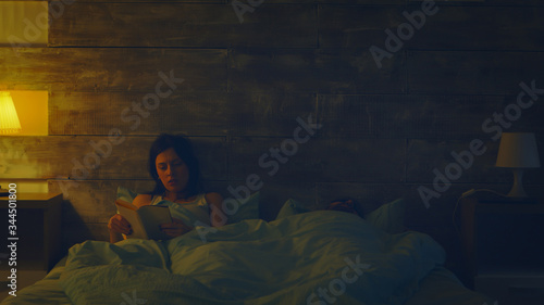 Beautiful wife reading a book in a dark room while his husband is sleeping. Lamp turned on.